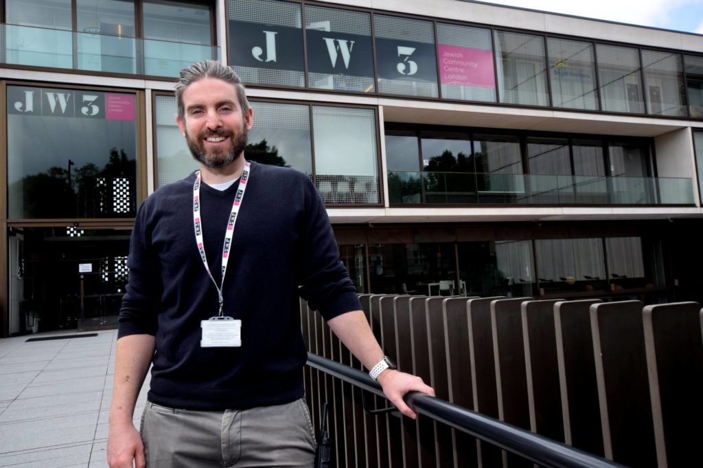 A white man wearing a lanyard stands in front of JW3 building
