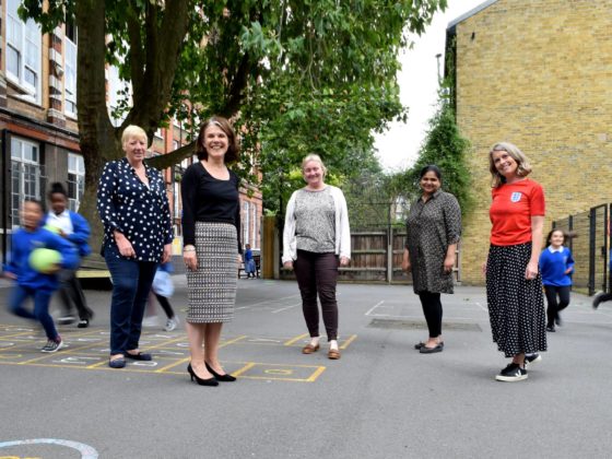 Five women stand in a school playground while children run past