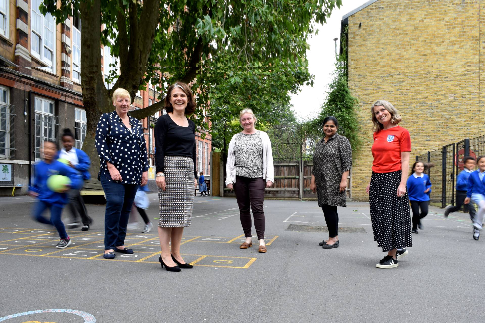 Five women stand in a school playground while children run past