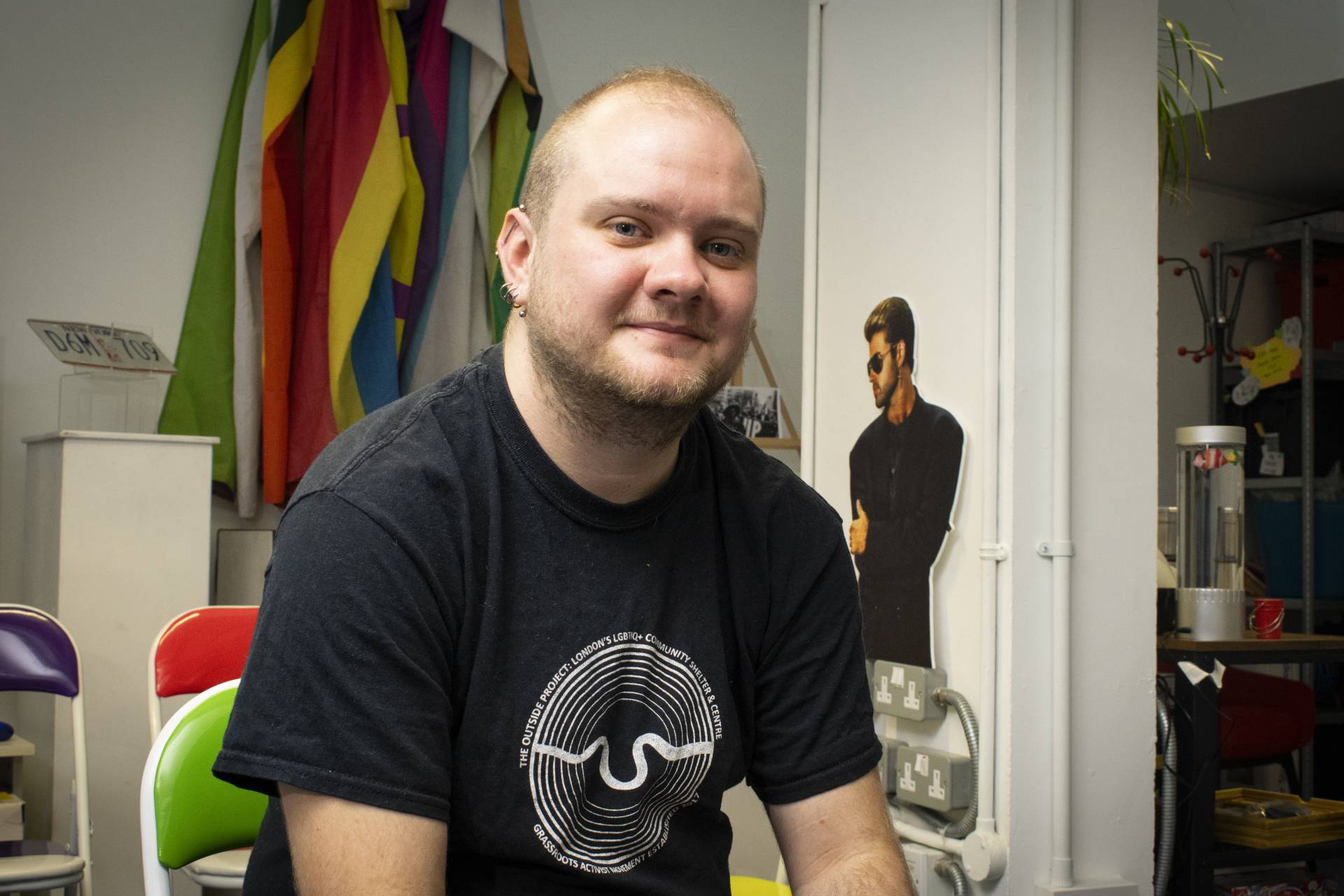 A white man with close cropped blonde hair and ear piercings, wearing a black T-shirt smiles for the camera