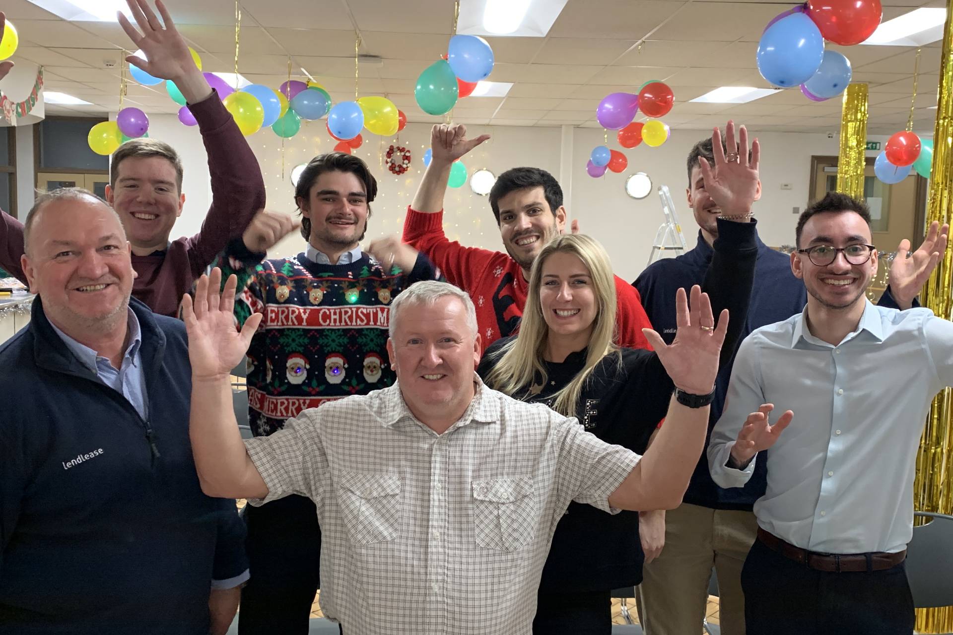 A group of people in an office decorated with ballons and Christmas decorations raise that hands and smile