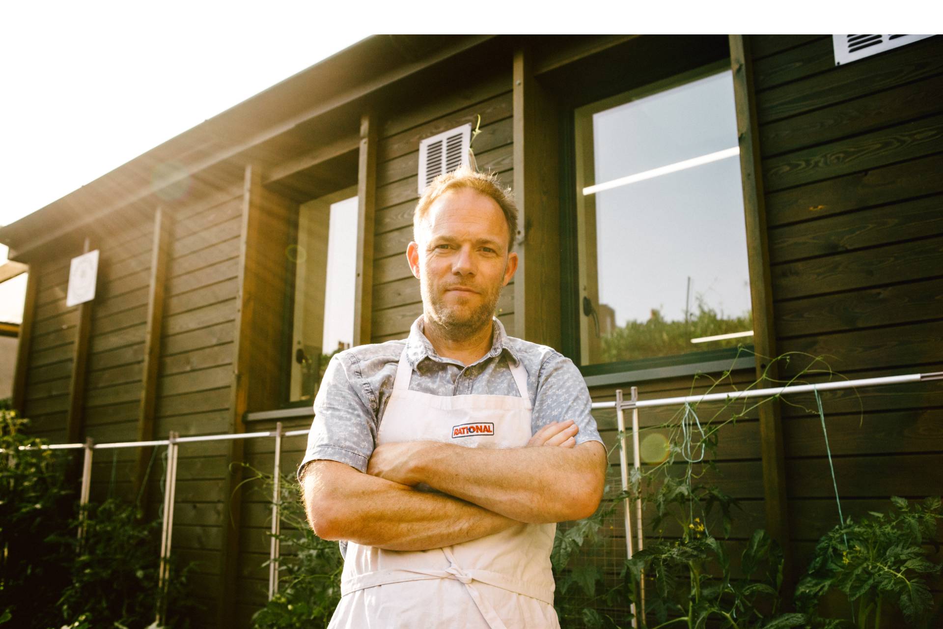 A white man wearing an apron and folded arms stands outside in the sunshine