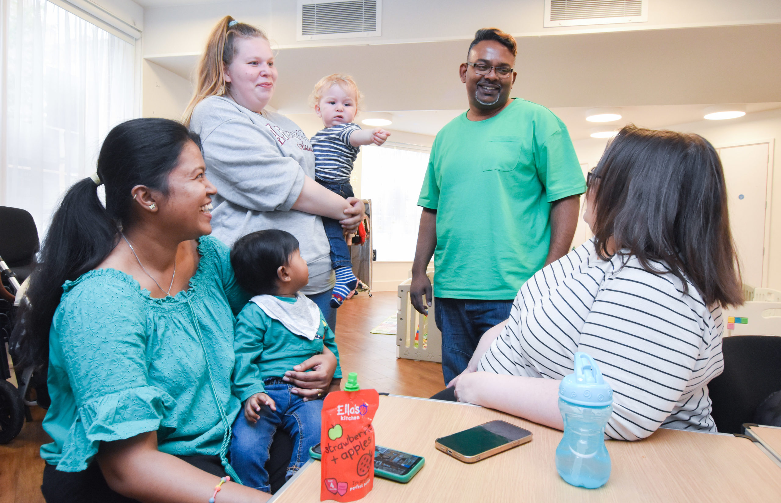 Mel tells a joke and three parents and their babies are laughing and smiling