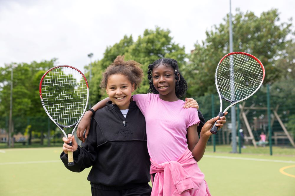 Two girls on a tennis court smiling and holding tennis rackets