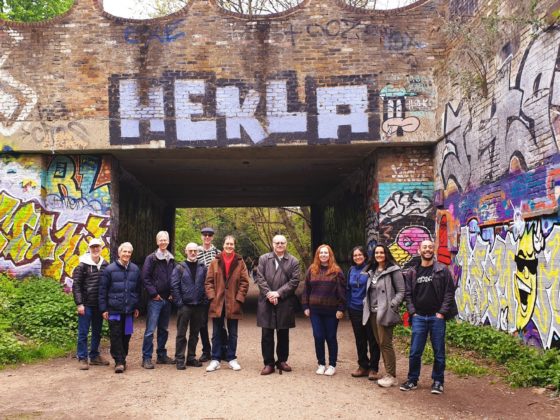 A group of 11 Forum+ members stand outside, in front of a graffiti wall on a walking trip.