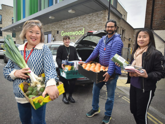 Sylvania and Arun stand with a young person and parent outside the Greenwood centre holding crates of fruit and vegetables. Everyone is smiling.