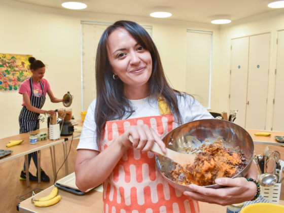 Charise stands inside the community centre at Bourne Estate smiling at the camera, wearing an apron and holding a mixing bowl of ingredients with a wooden spoon.