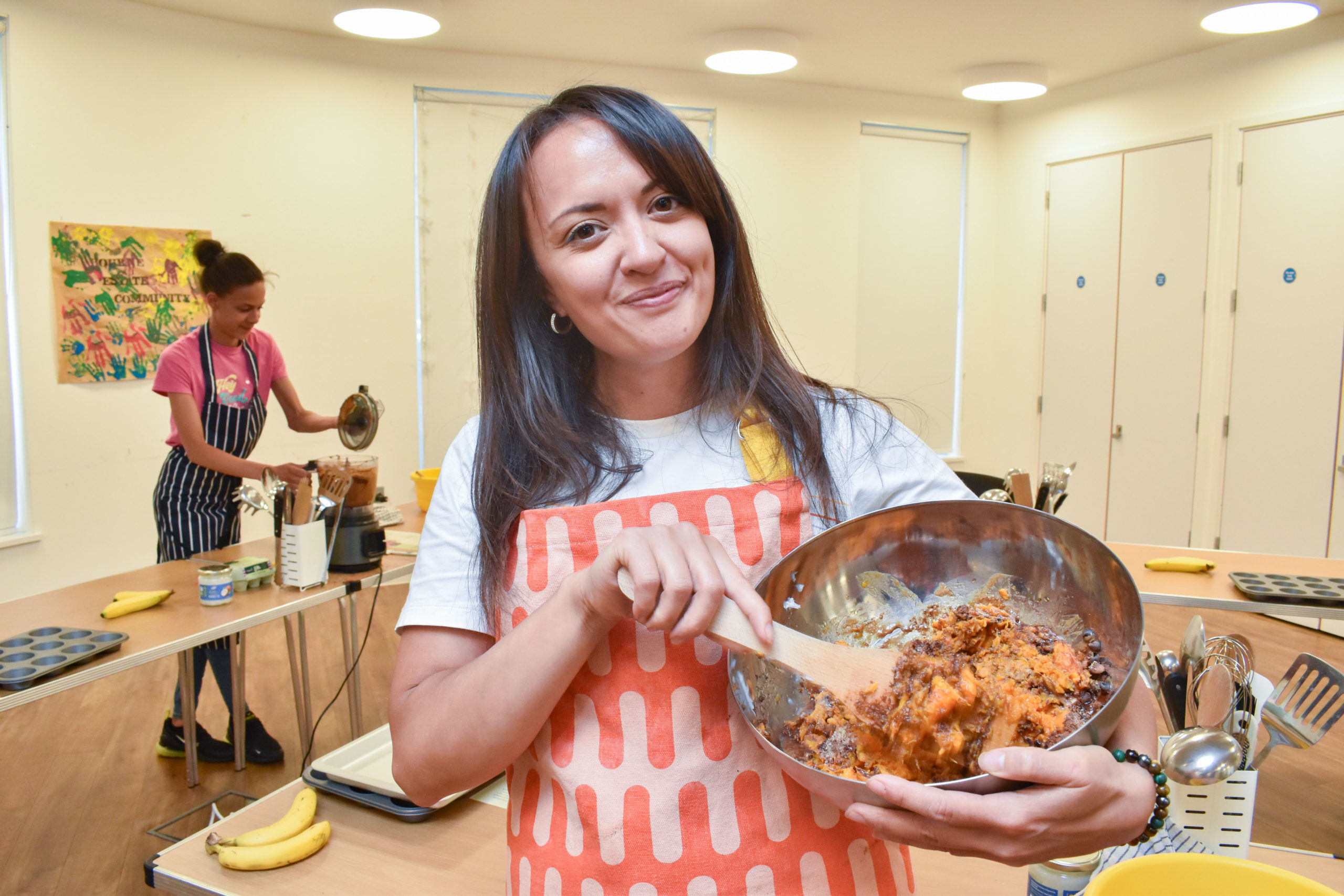 Charise stands inside the community centre at Bourne Estate smiling at the camera, wearing an apron and holding a mixing bowl of ingredients with a wooden spoon.