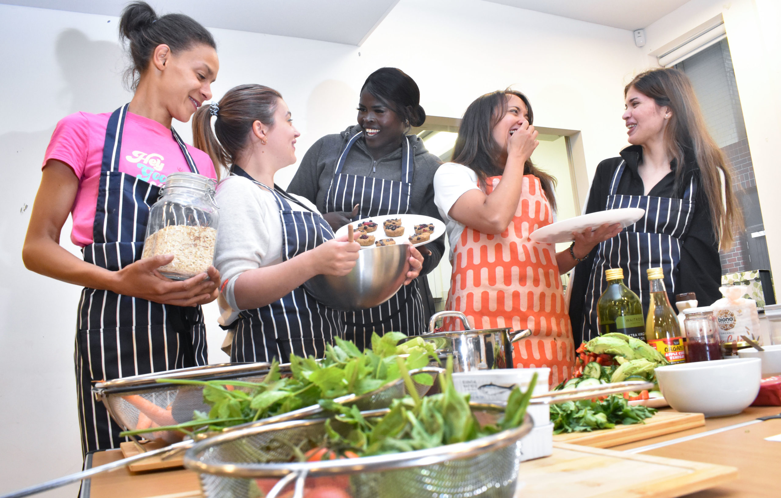 A group of four female workshop participations stand together, with Charise, laughing with each other. There is fresh salad at the front of the photo and each woman is holding a plate of healthy snacks they have made.