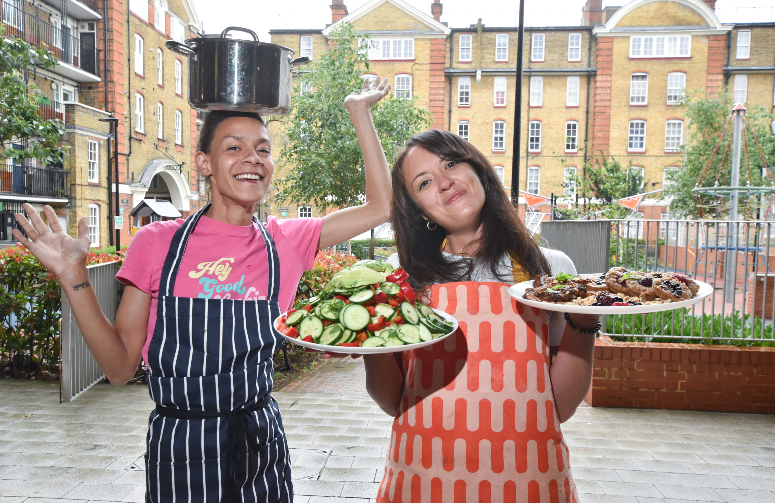 Charise and a female workshop participant are standing outside at the Bourne Estate. Both are wearing aprons. Charise is holding two large plates of fresh salad and sweet potato brownies. The participant is smiling at the camera while balancing a saucepan on her head.