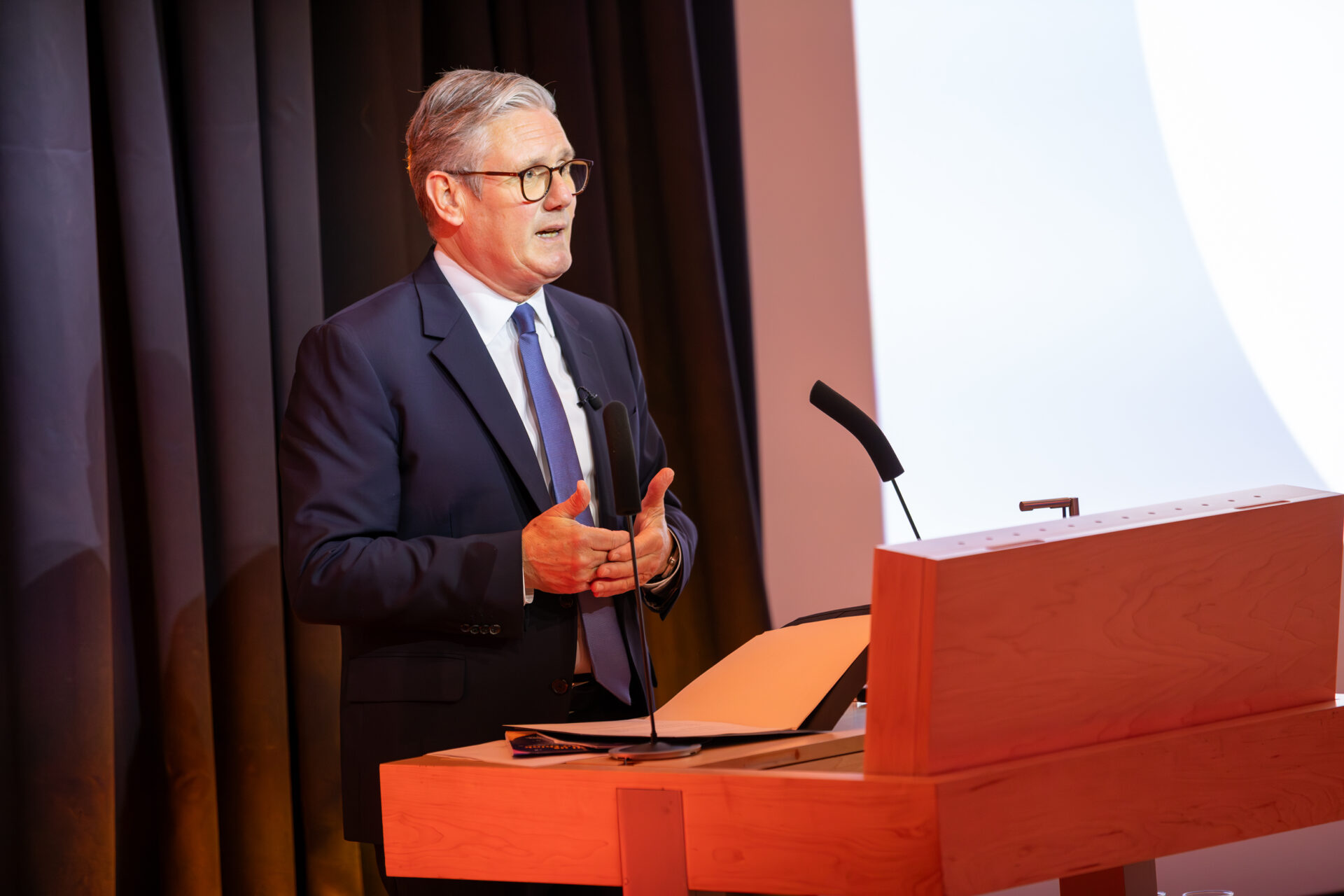 Sir Kier Starmer, Prime Minister and MP for Holborn and St Pancras, stands at a podium with two microphones. He is mid-speech, addressing the audience. He is wearing a navy blue suit, blue tie, white shirt and glasses.