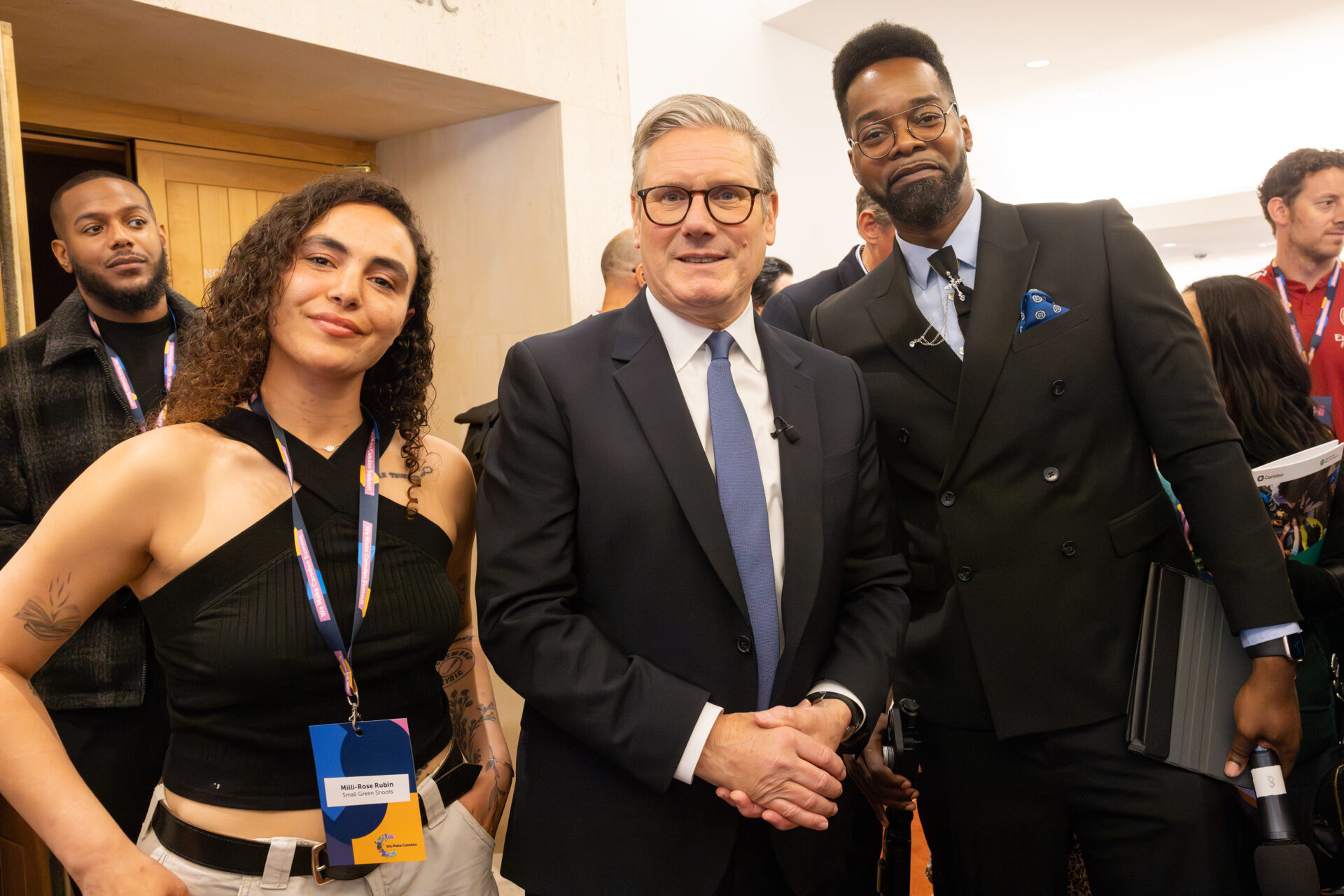Sir Kier Starmer stands between Milli-Rose and Alex Emeh-Gapo (event hosts) - they are all smiling. Milli-Rose has curly hair and wears and strappy black top and cream combat trousers. Alex wears a black suit, blue shirt and navy tie. Keir Starmer wears a navy suit, blue tie, white shirt and glasses. There are other people in the background of the photo.