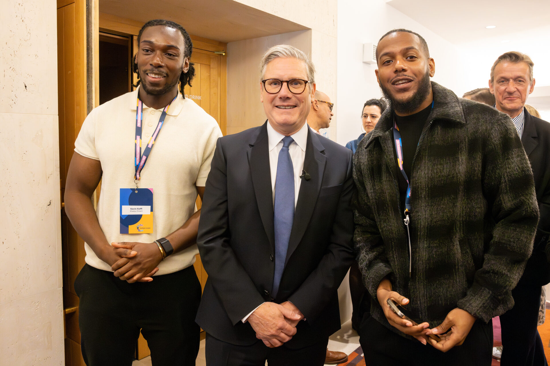 Sir Kier Starmer stands between Kevin Koffi and Jadye Allen from Khadys Dream - everyone is smiling. Kevin wears a cream t-shirt and We Make Camden Lanyard. Kier wears a navy suit, blue tie, white shirt and glasses. Jayde wears a black and green checked jacket and a We Make Camden lanyard. Cllr Richard Olszewski is in the background.