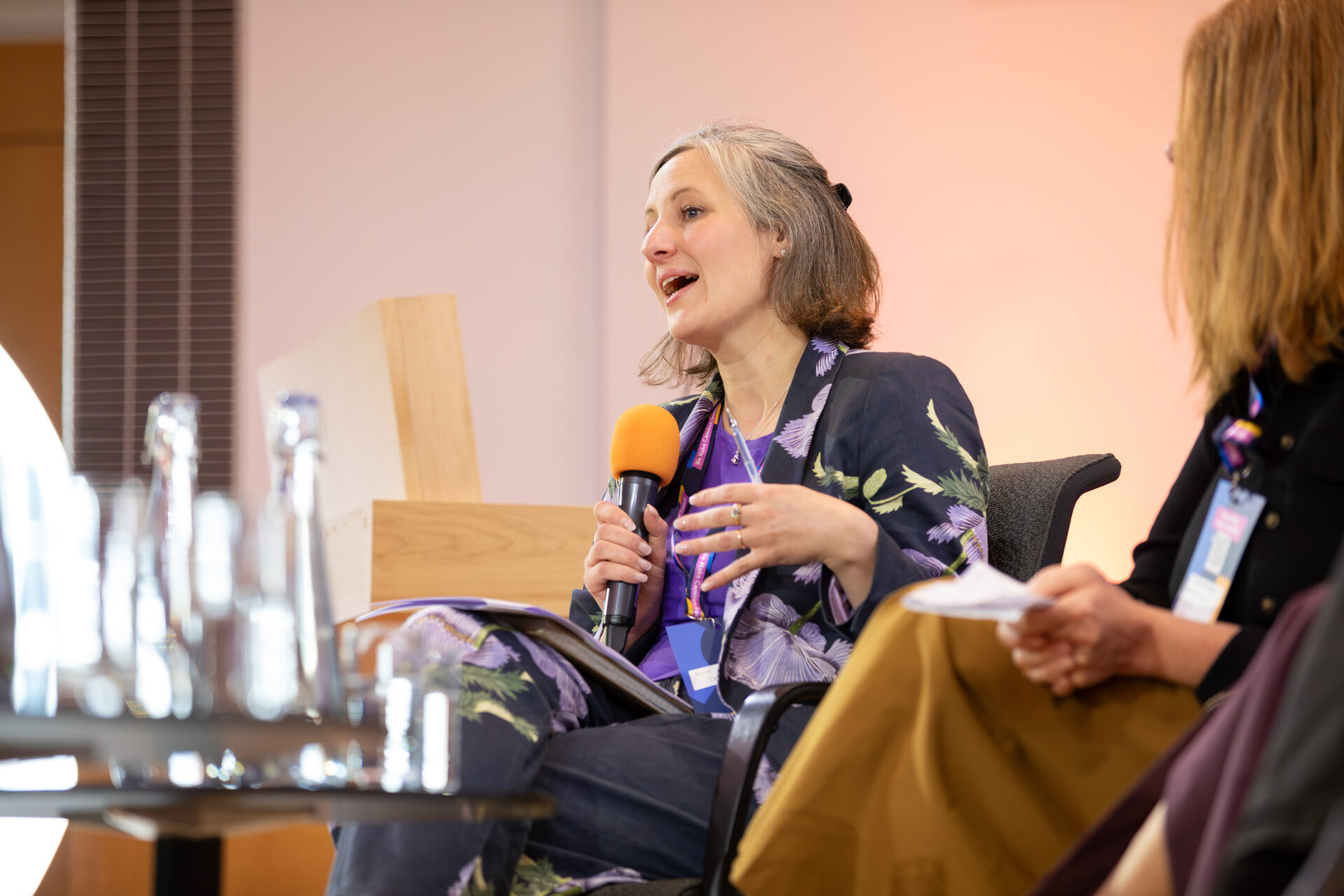 A member of the Child Poverty Panel is speaking on stage. She is sat in a chair holding a black and orange microphone. She wears a navy floral printed blazer with purple flowers on.