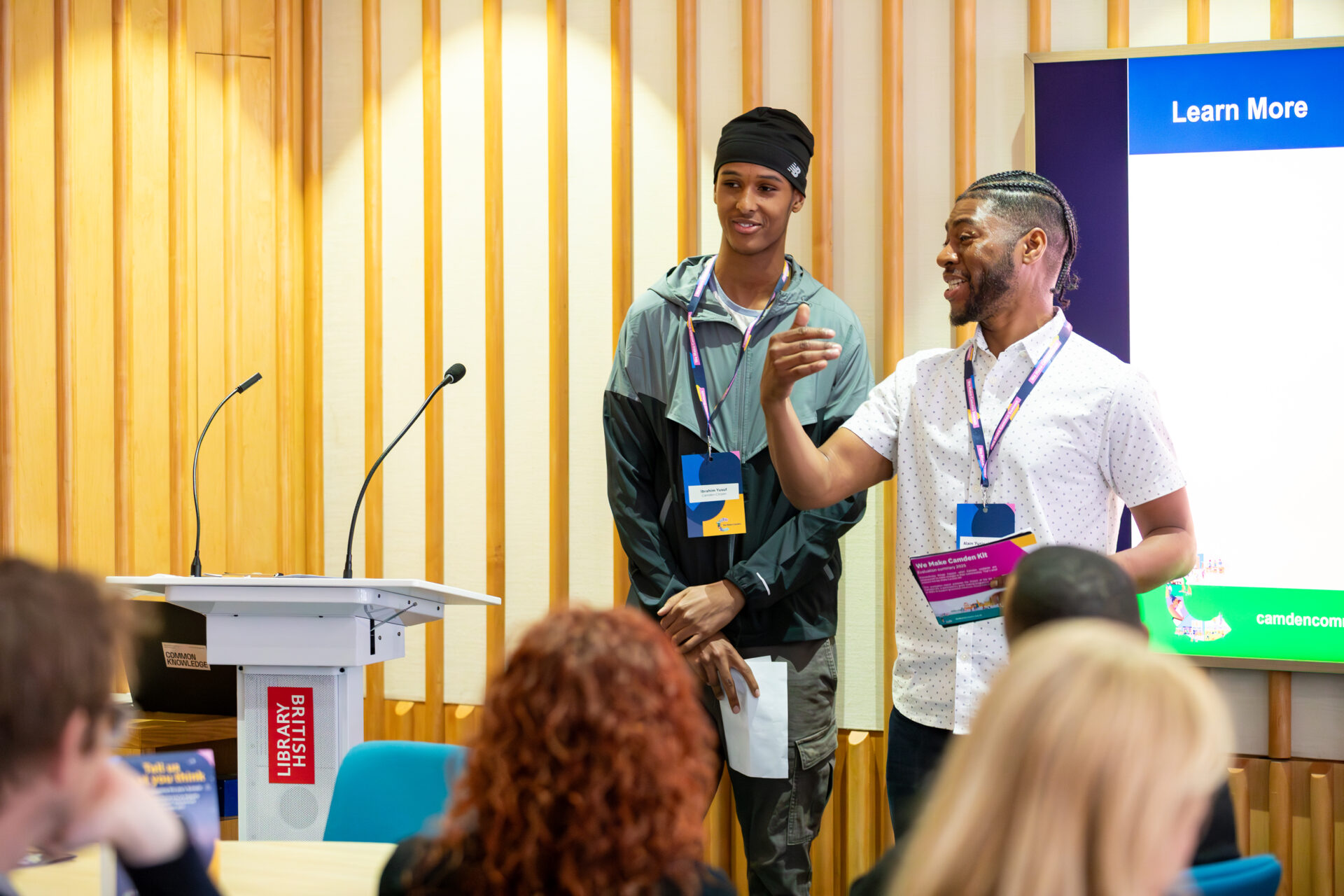 Alex and a member of the Camden Community Wealth Fund Youth Panel stand in front of a podium with microphones, presenting to the audience. Both wear We Make Camden Summit lanyards and stand in wood panelled room.