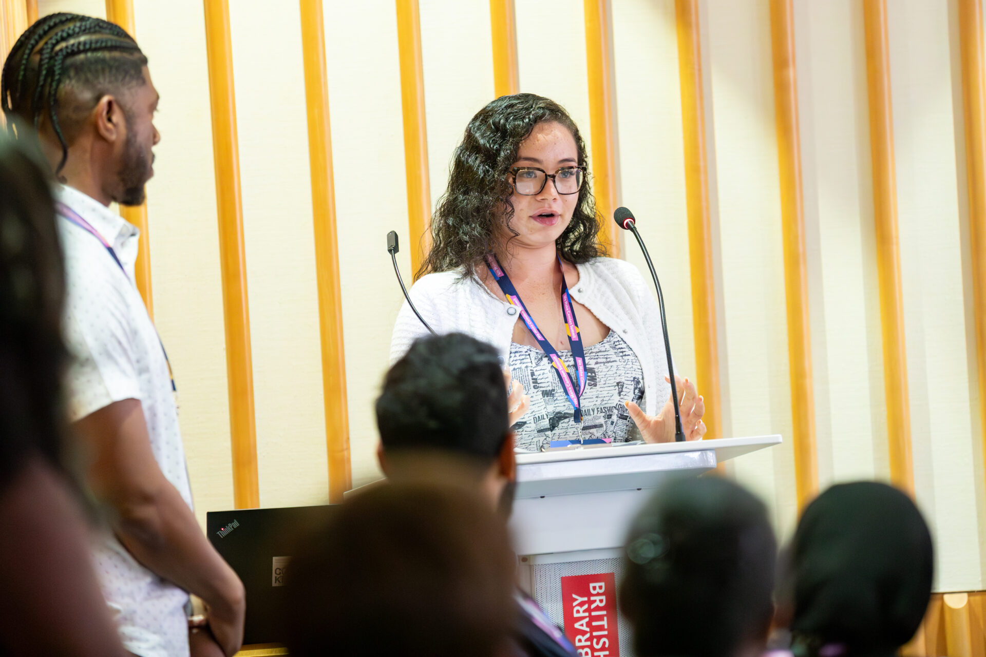 A member of the Camden Community Wealth Fund Youth Panel is standing at a British Library podium addressing a room of people. She is wearing a black and white top, white cardigan, We Make Camden lanyard and glasses. There is wooden panelling on the walls behind and Alain 'Fusion' Clapham can be seen standing to the side of the room - he wears a white short sleeved shirt.