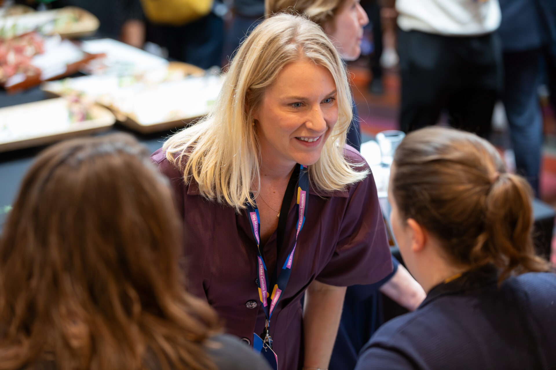 A woman from the Council's Public Health team bends down to speak to a guest. She is wearing a purple dress and a We Make Camden lanyard - she is smiling. The face of the guest she is speaking to is blurred.