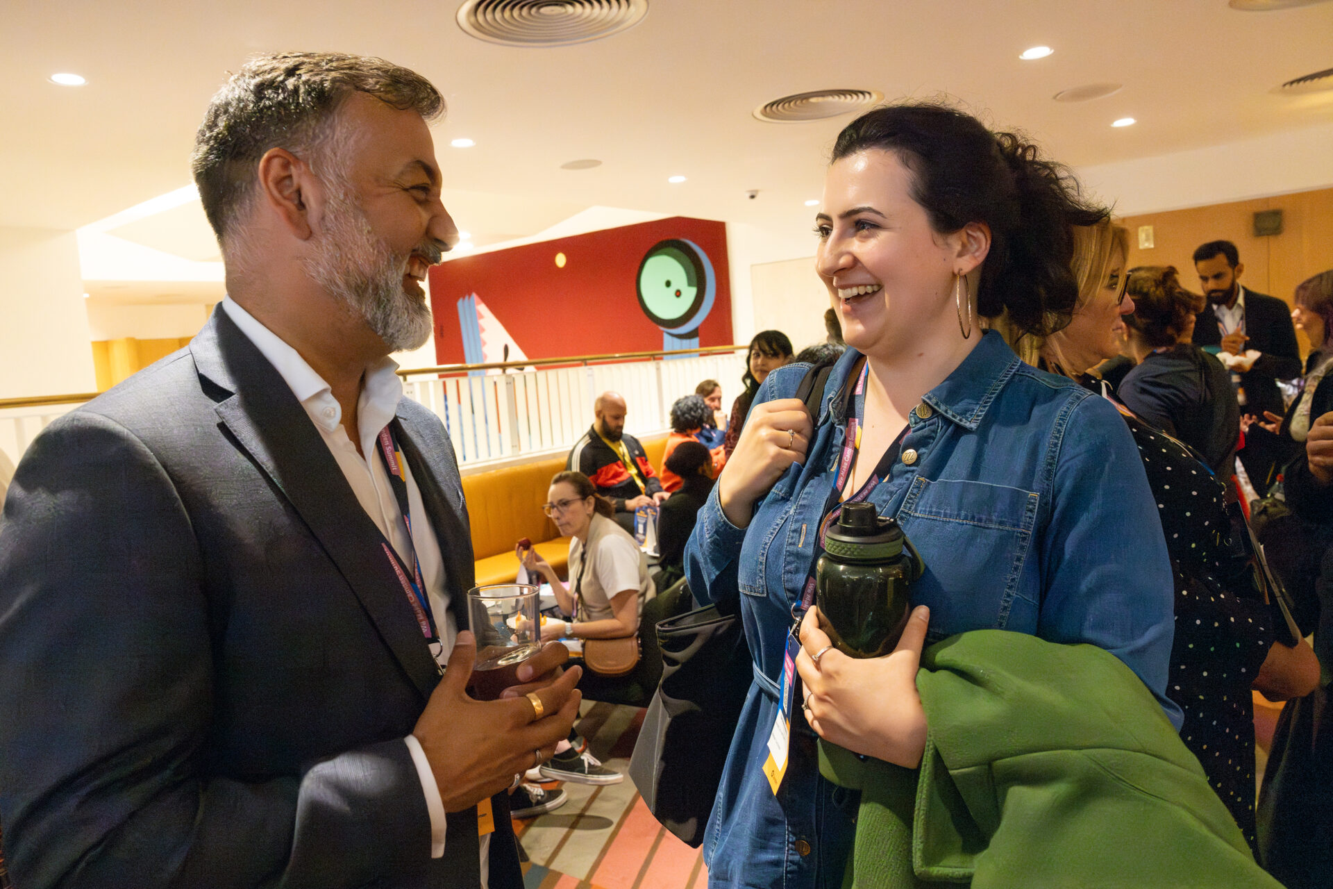 Two event guests are in conversation and laughing. The man wears a grey suit, white shirt and We Make Camden Summit lanyard, and the woman wears a blue denim jumpsuit, gold hoops and a lanyard - she also holds a water bottle and has a green coat over her arm. Other guests are in the background of the photo.