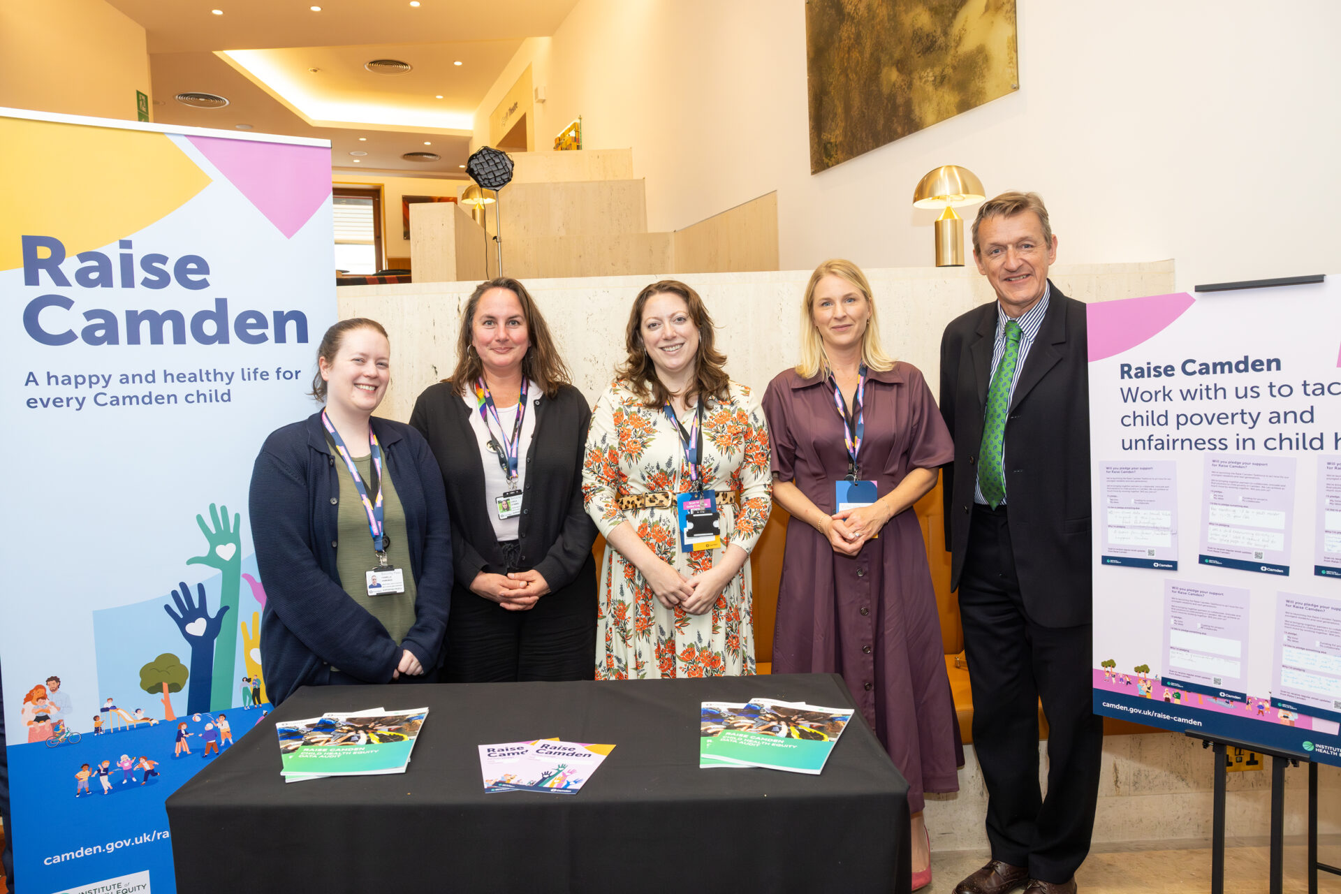Three women from Camden Council's Public Health team stand with the Leader of Camden Council, Cllr Richard Olszewski, with a table in front of them which has the new Raise Camden report on. Beside them is a Raise Camden pull-up banner and a board asking guests to make to tackle child poverty.