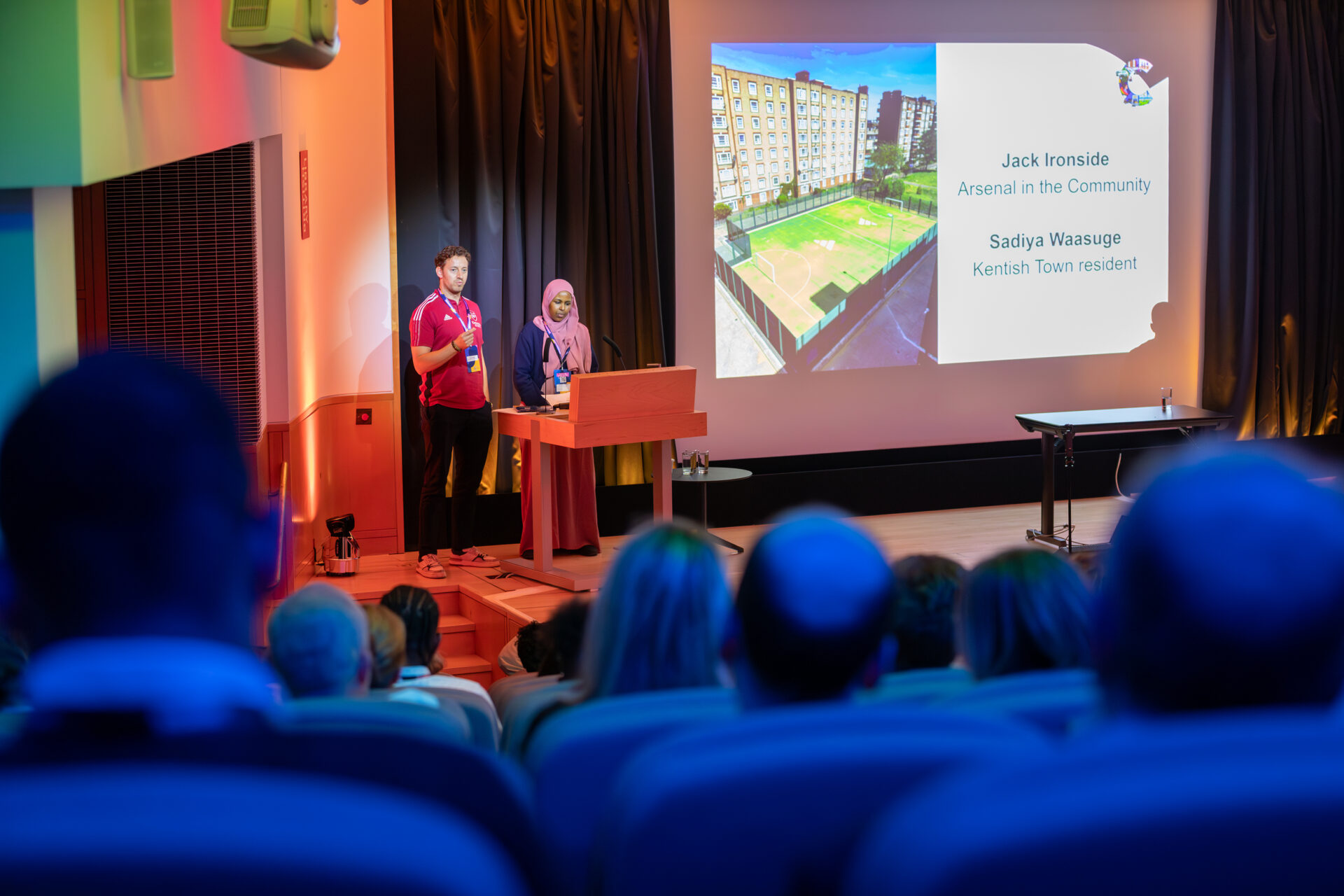 Jack and Sadiya stand at a podium in a lecture theatre, addressing the audience. Jack is wearing a red Arsenal sports shirt and Sadiya is wearing a pink headscarf and navy jumper. Behind them, a large screen displays an image of the new sports pitch and reads: 'Jack Ironside – Arsenal in the Community' and 'Sadiya Waasuge – Kentish Town resident.'