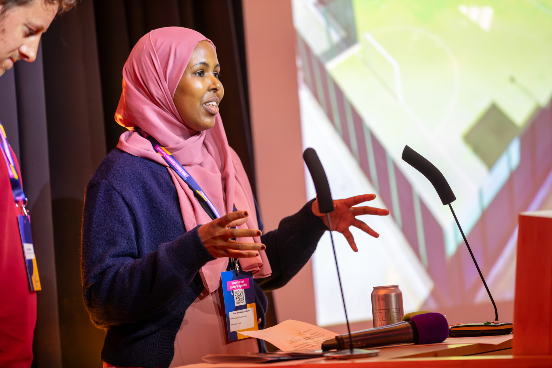 Sadiya wears a pink headscarf and a blue jumper and stands at a podium with two microphones, gesturing while speaking. Jack is partially visible on the left. In the background, a large screen displays an aerial view of the Peckwater Estate sports pitch.