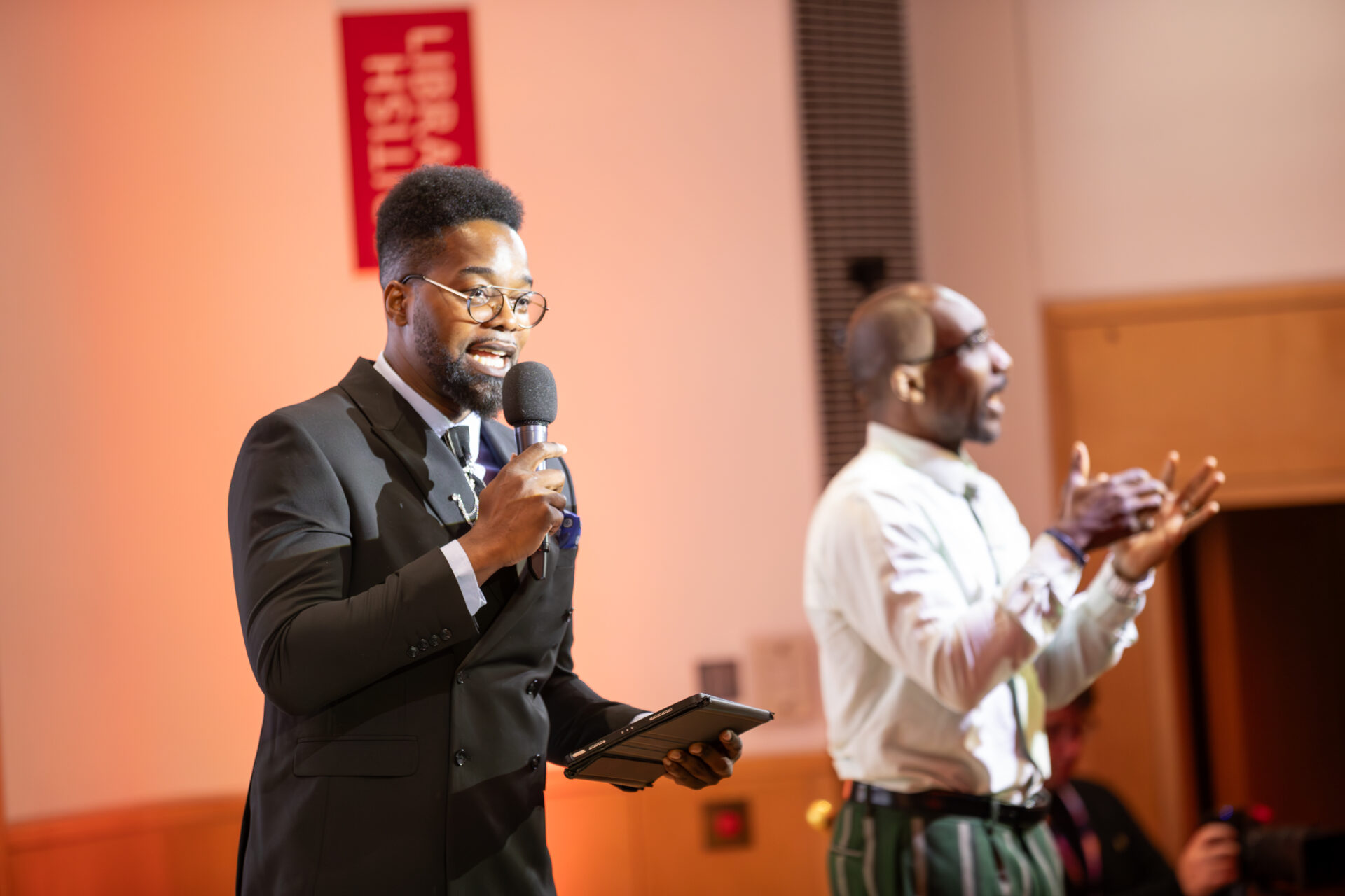 Alex and a BSL sign language interpreter are on stage at the event. Alex is dressed in a black suit, holds a microphone and a tablet. The interpreter is in a white shirt and green striped trousers signing. A red vertical sign in the background reads 'British Library'