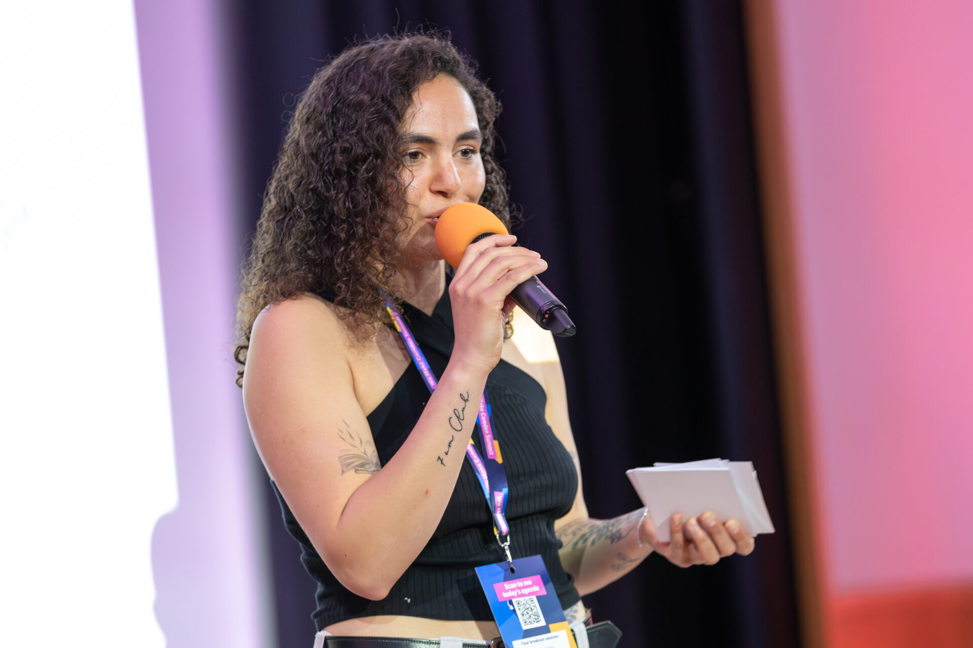 Milli-Rose is speaking into a microphone while holding index cards. She is wearing a black sleeveless top and a Summit lanyard on. A white screen and dark curtains are in the background.