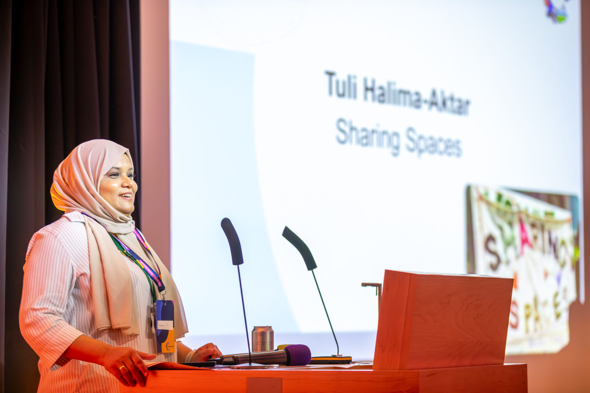 Tuli is a light-colored hijab and a white shirt. She stands at a podium with two microphones, delivering a presentation. Behind her, a projection screen displays the text 'Tuli Halima-Aktar Sharing Spaces' alongside artwork featuring the same phrase.