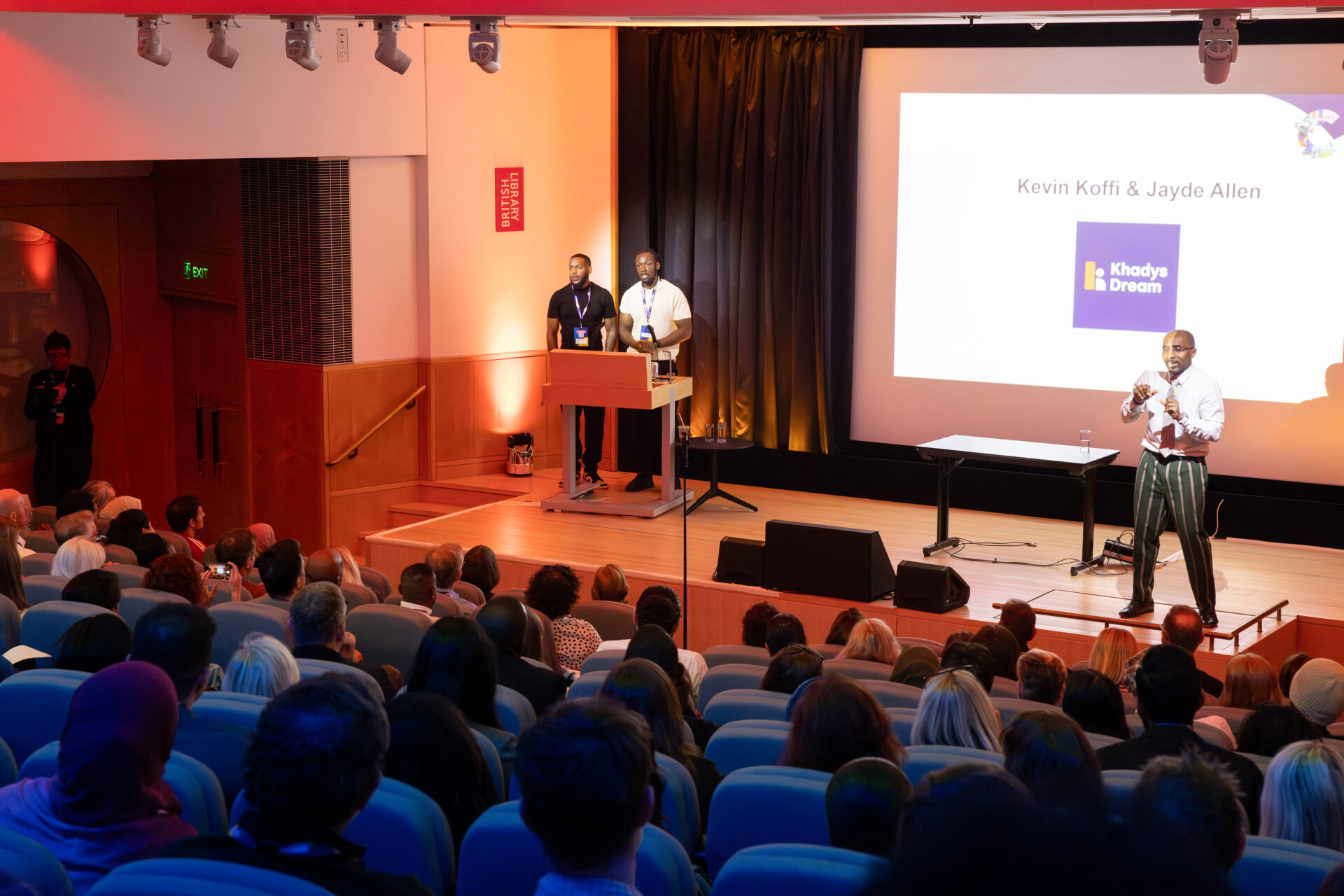 Kevin Koffi and Jayde Allen are on the stage facing towards the audience sitting in the auditorium. A white slide is on a large screen behind them, reading 'Kevin Koffi & Jayde Allen' alongside the blue Khadys Dream logo. There is a British sign language interpreter on the right of the stage.