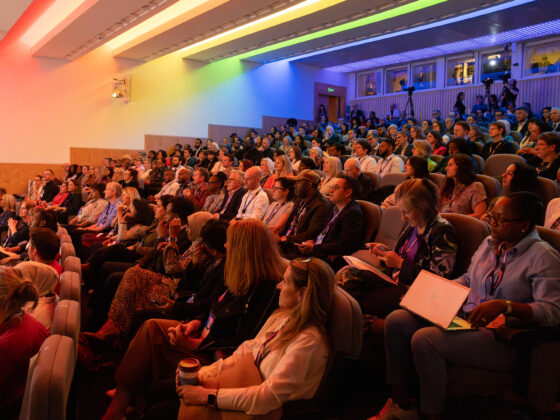 A large number of guests at the Summit sit in a auditorium facing the stage. There are rainbow coloured lights in the ceiling, creating a colour hue over guests.