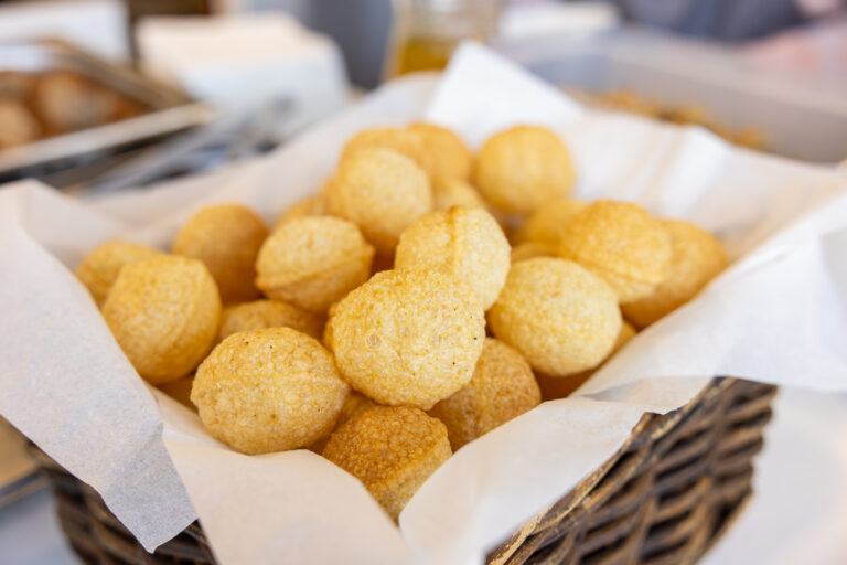 A basket lined with white parchment paper, filled with round, golden-brown bread rolls or puffs, likely freshly baked.