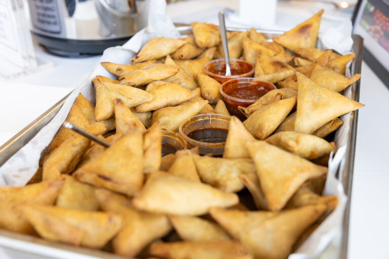 A large tray of golden-brown triangular samosas, accompanied by three small containers of dipping sauces—dark brown, reddish-brown, and orange-red - each with a serving spoon, set against a blurred kitchen background.