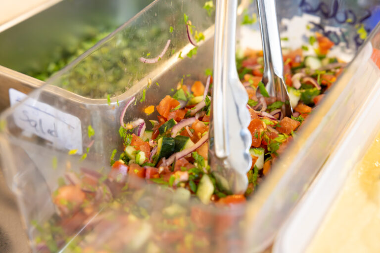 A container of fresh salad with diced tomatoes, cucumbers, red onions, and herbs, served with metal tongs.
