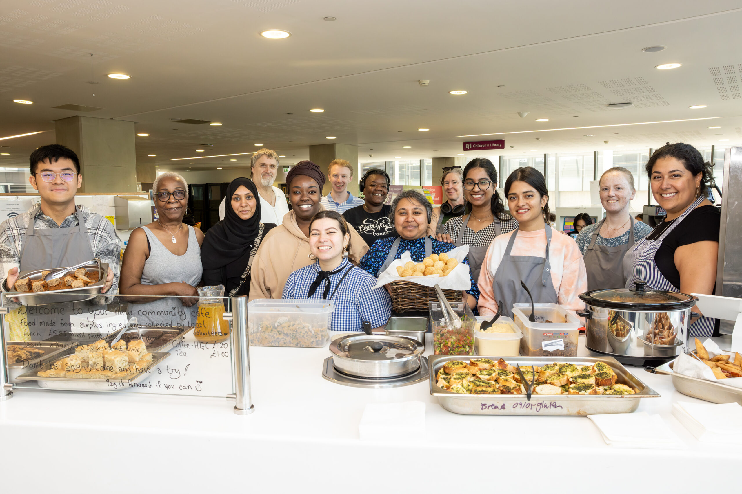 A group of smiling staff and volunteers standing behind a counter at the community café, with trays of food displayed in front of them.