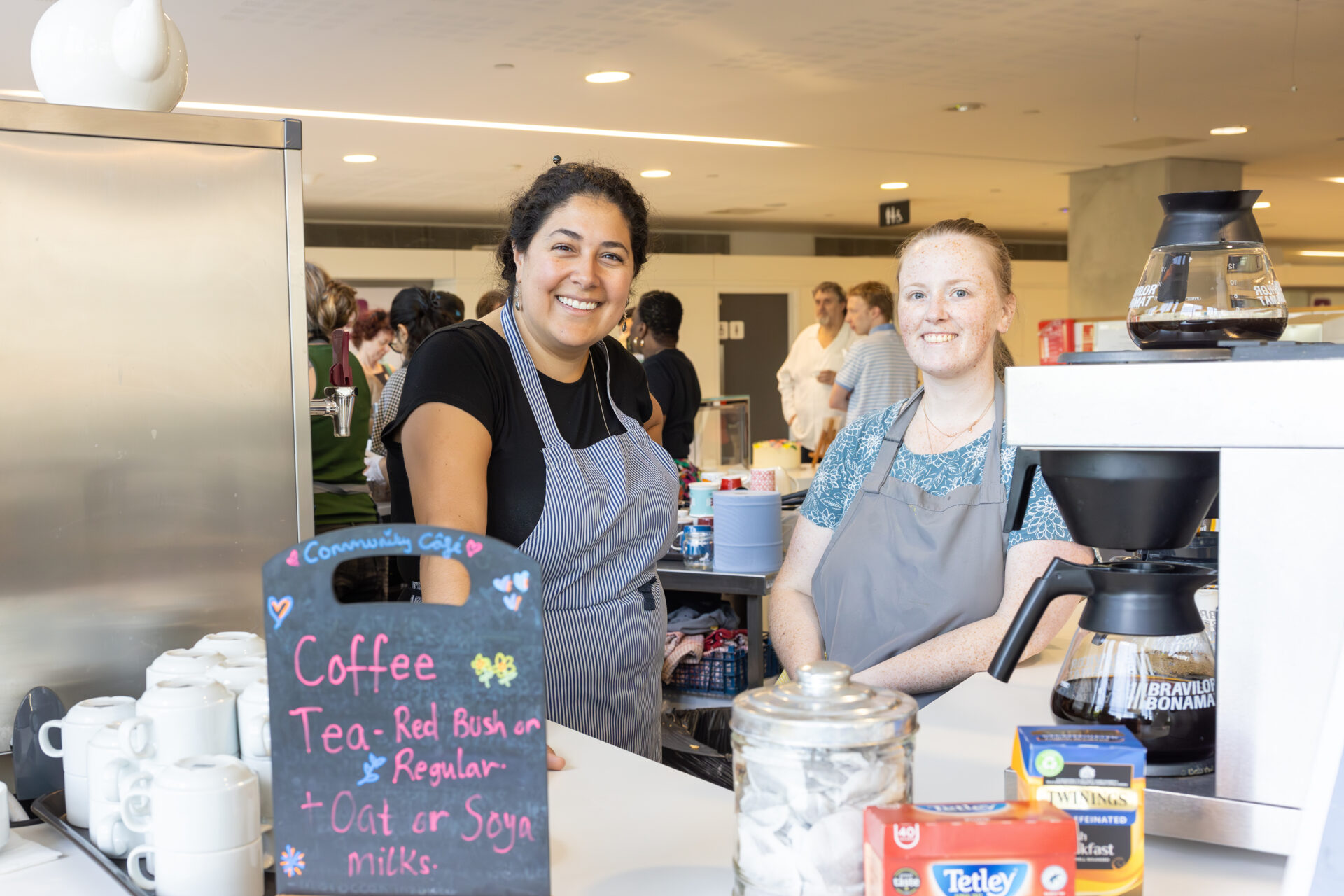 Two individuals behind a counter with coffee-making equipment and mugs. A chalkboard sign lists beverages including coffee, tea (Red Bush or Regular), and oat or soya milk options.