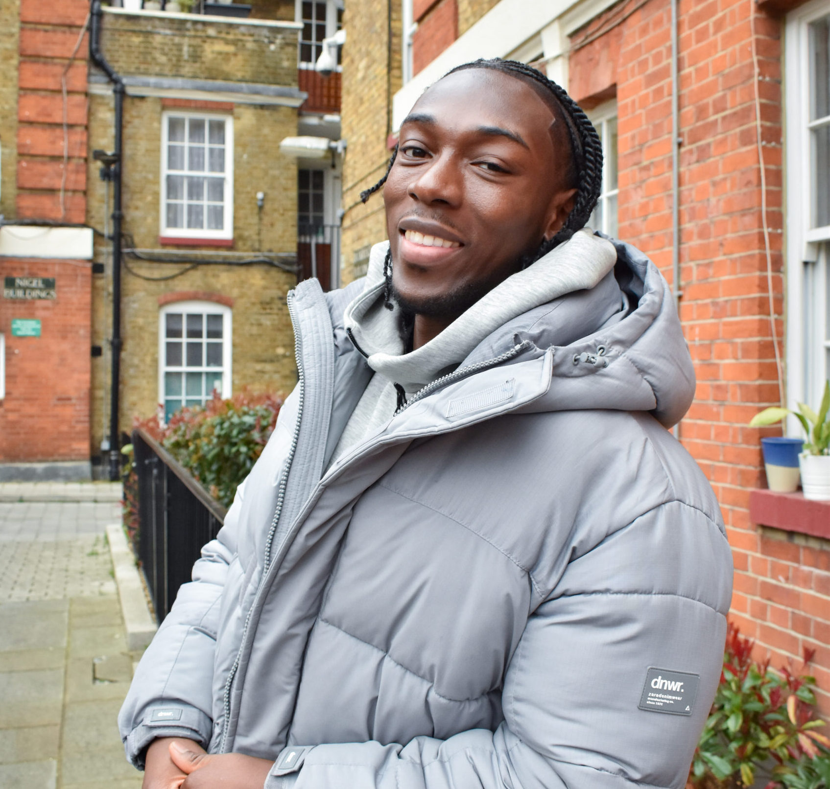 Kevin sands in front of a block of flats. He is leaning against a black metal fence, wearing a grey coat and is smiling at the camera.