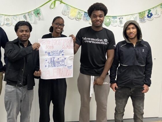 A group of young people standing together indoors, holding a handmade sign advertising free food and a local celebration.