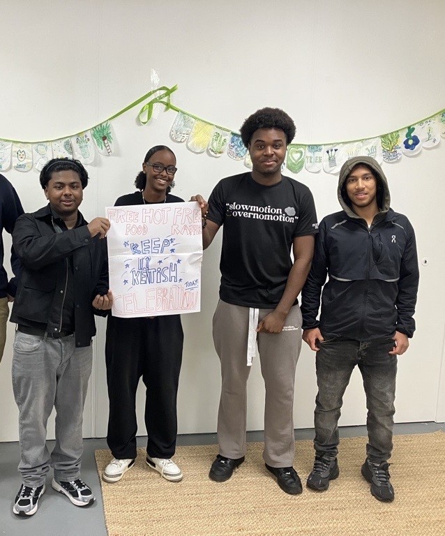 A group of young people standing together indoors, holding a handmade sign advertising free food and a local celebration.