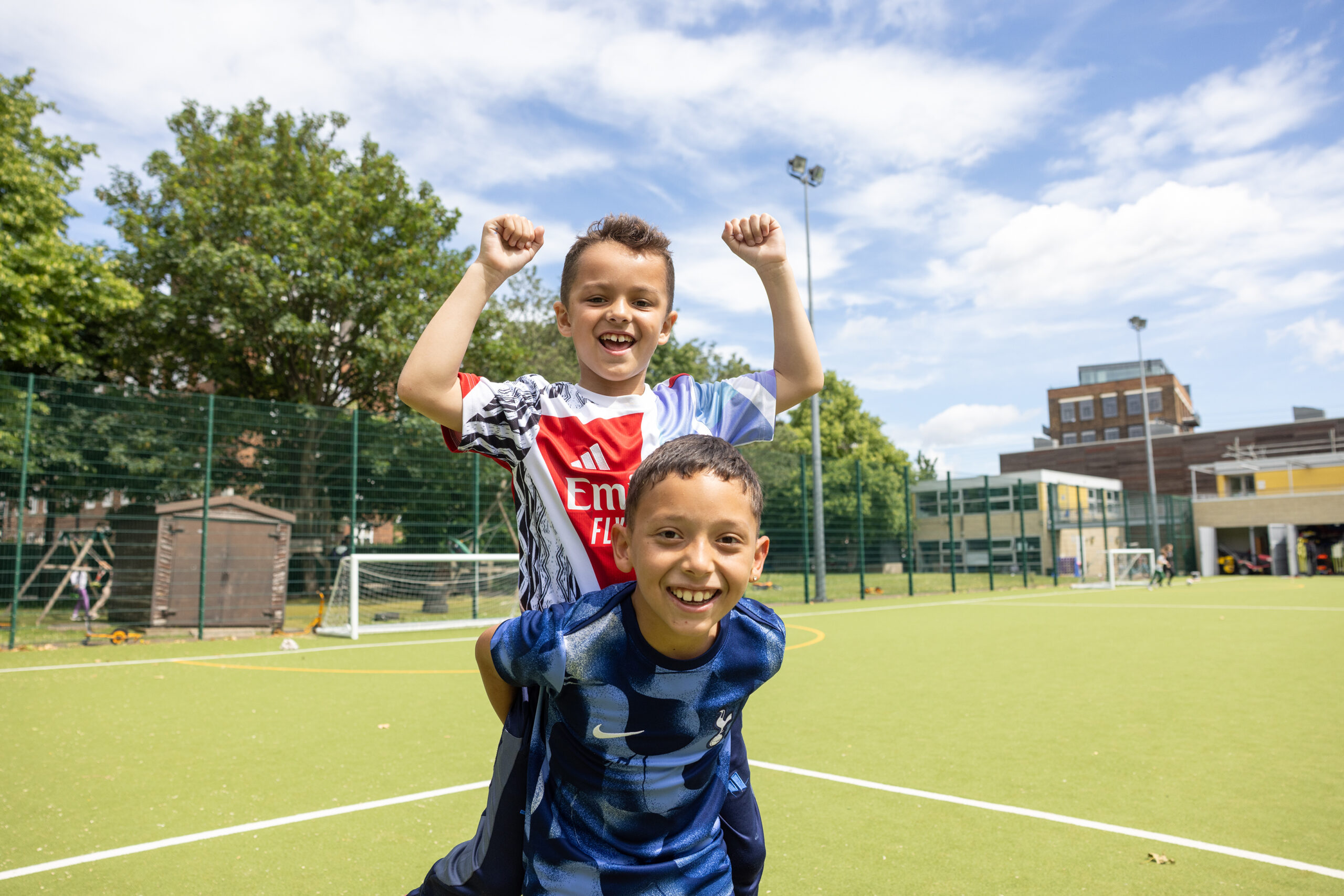 A boy is giving a younger boy a piggyback on a football pitch, as they both cheer to the camera.