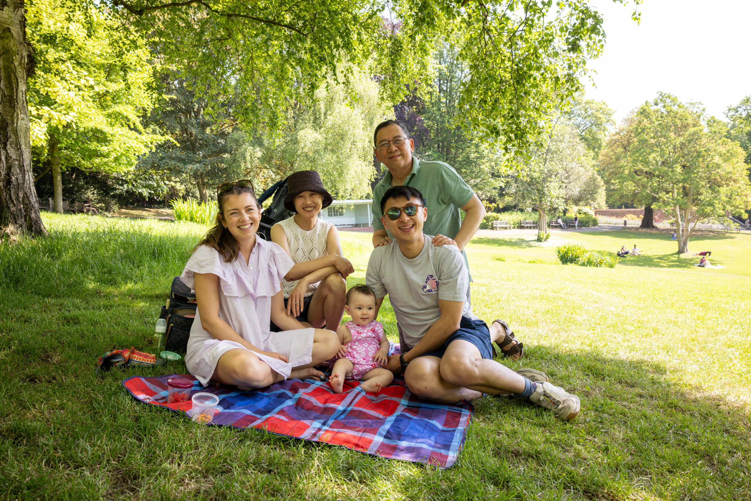 Four adults and a baby are sitting together on a blanket in the park, smiling at the camera.